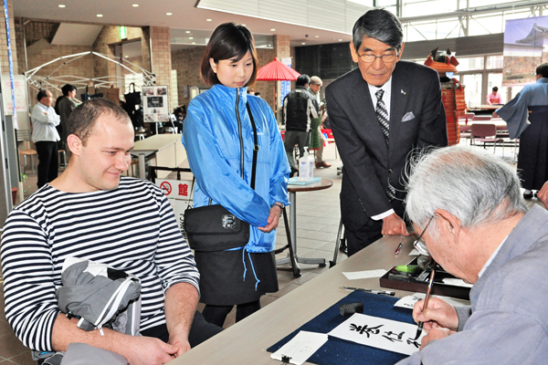 京都北都信用金庫舞鶴中央支店の金巻永理さん(中央左)や森屋理事長(同右)が助言し、漢字で名前を書いてもらう外国人観光客。(舞鶴市西市民プラザ)