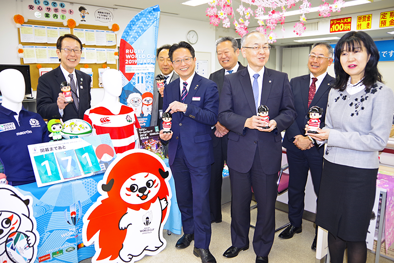 女性職員の手作りジャージを着た「信ちゃん貯金箱」を手に栗田区長(右)を出迎える守田氏(左から5人目)ら(4月2日、新横浜支店)