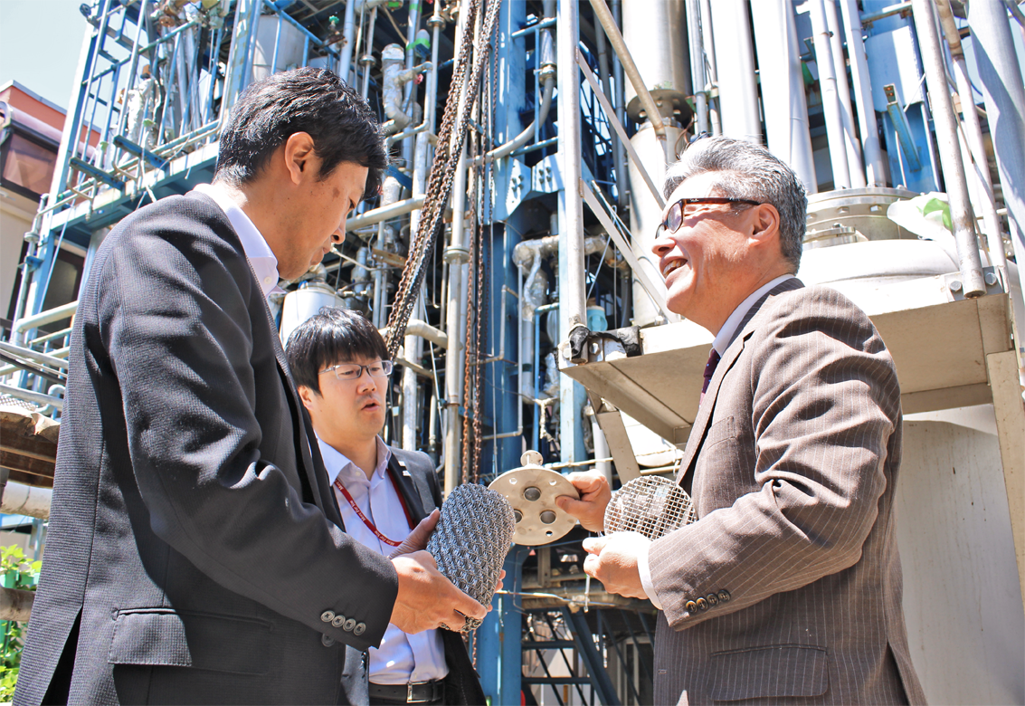 こまめに経営状況を把握するため、取引先のトウトクエンジ大田二郎社長(右)から同社蒸留塔の前で製品の説明を受ける橋口剛洋支店長(左)と須賀光一営業課長(5月8日、トウトクエンジ三郷工場)