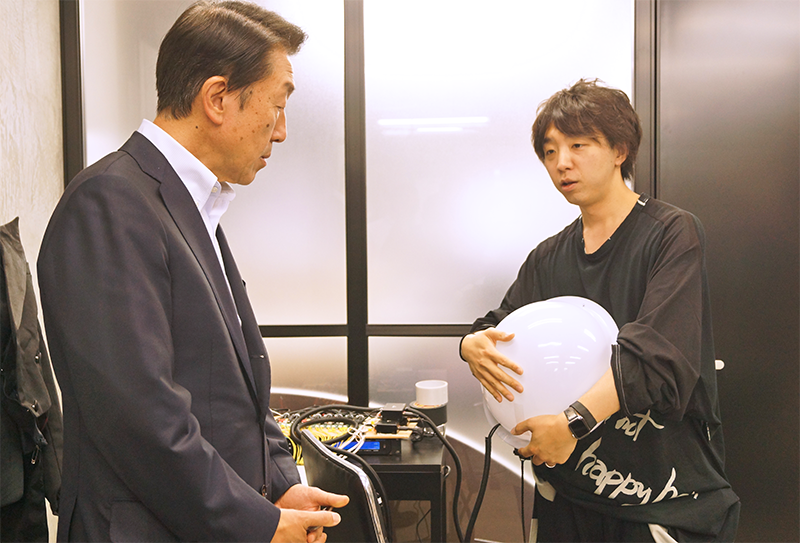 聴覚障がい者も波動で音楽を体感できる球体型デバイスの説明を落合氏から聞く関根社長(左、6月4日、東京・神田)