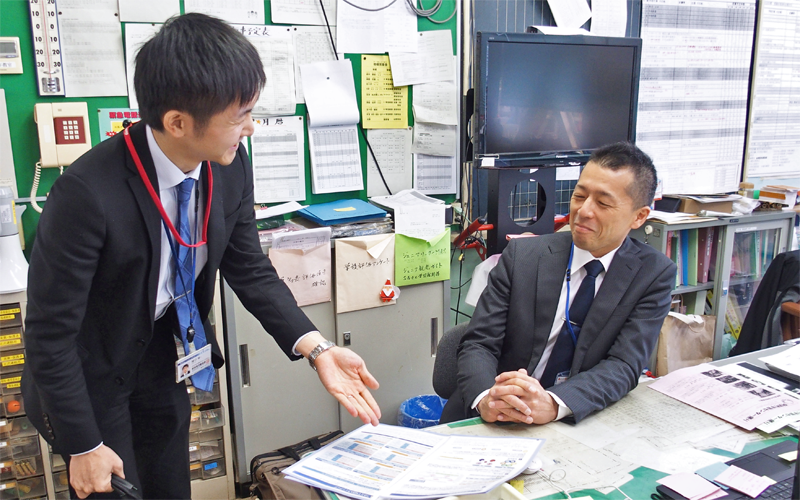 阿賀町立三川小学校の田中一史校長(右)に職員室で商品説明をする樋口裕主任(1月9日)