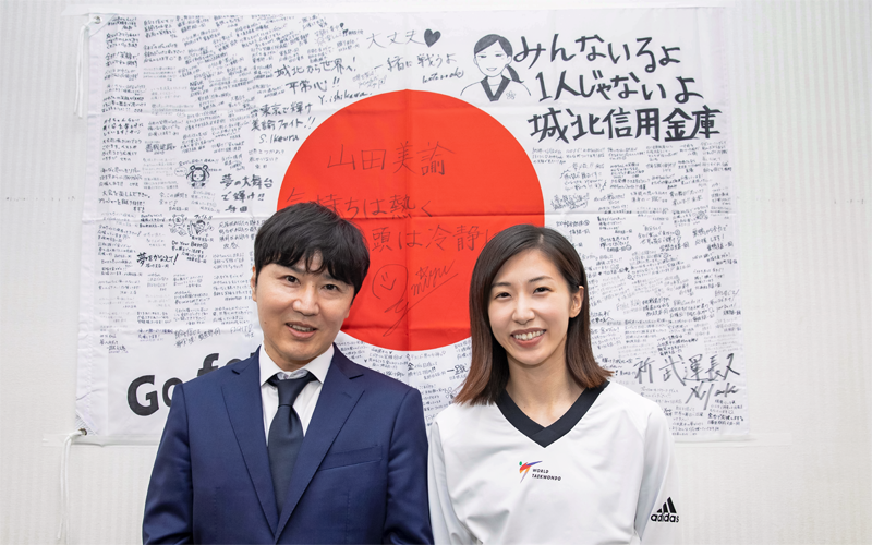役職員の寄せ書きを添えた日本国旗の前に立つ城北信金の大前理事長(左)と山田選手(7月7日、本部)
