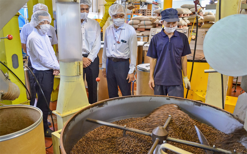 ダートコーヒーの中西泰仁業務部長（右から2人目）からコーヒー豆の焙煎工程などのレクチャーを受ける北川昌志支店長（同3人目）と小林義和支店長代理（左、7月16日、ダートコーヒー白山工場）