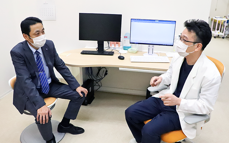 診察室でみずの内科・外科クリニックの水野史人院長(右)から近況を聞く、成長戦略部の八代副調査役
