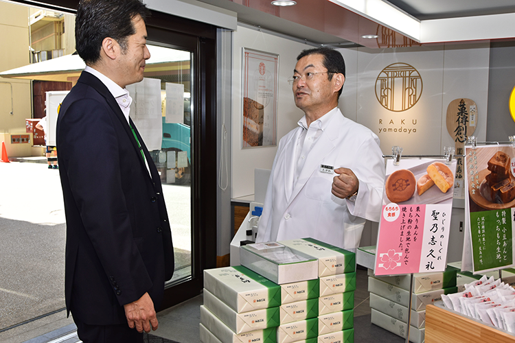 もみじ饅頭の製造販売を手掛けるやまだ屋の田川繁章取締役(右)と談笑する岩本支店長(5月17日、廿日市市宮島町)