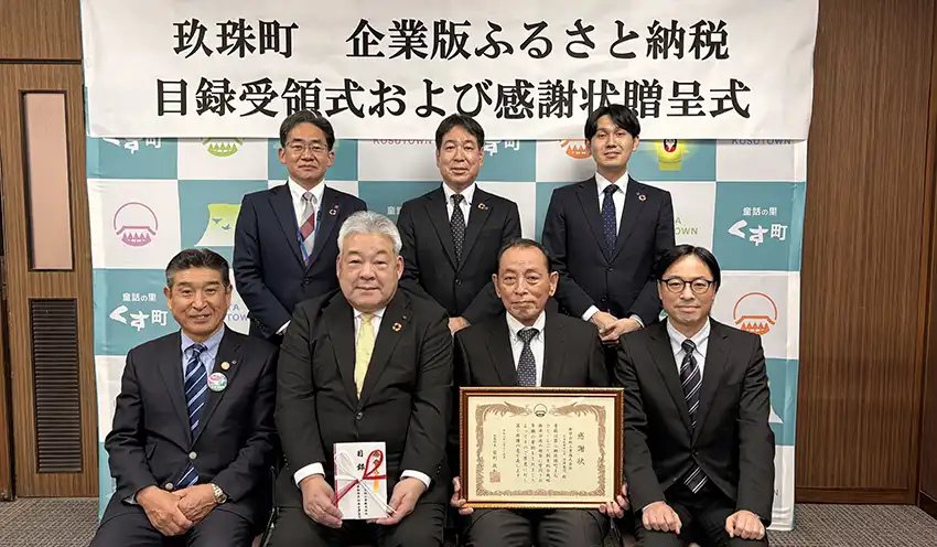 寄付目録を持つ宿利政和・玖珠町長（前列左から2人目）と感謝状を掲げる古澤憲司・新栄合板工業社長（同3人目）。後列左から、大分銀行地域創造部の吉田智之部長、梅尾正和支店長、担当者（3月25日、玖珠町役場）
